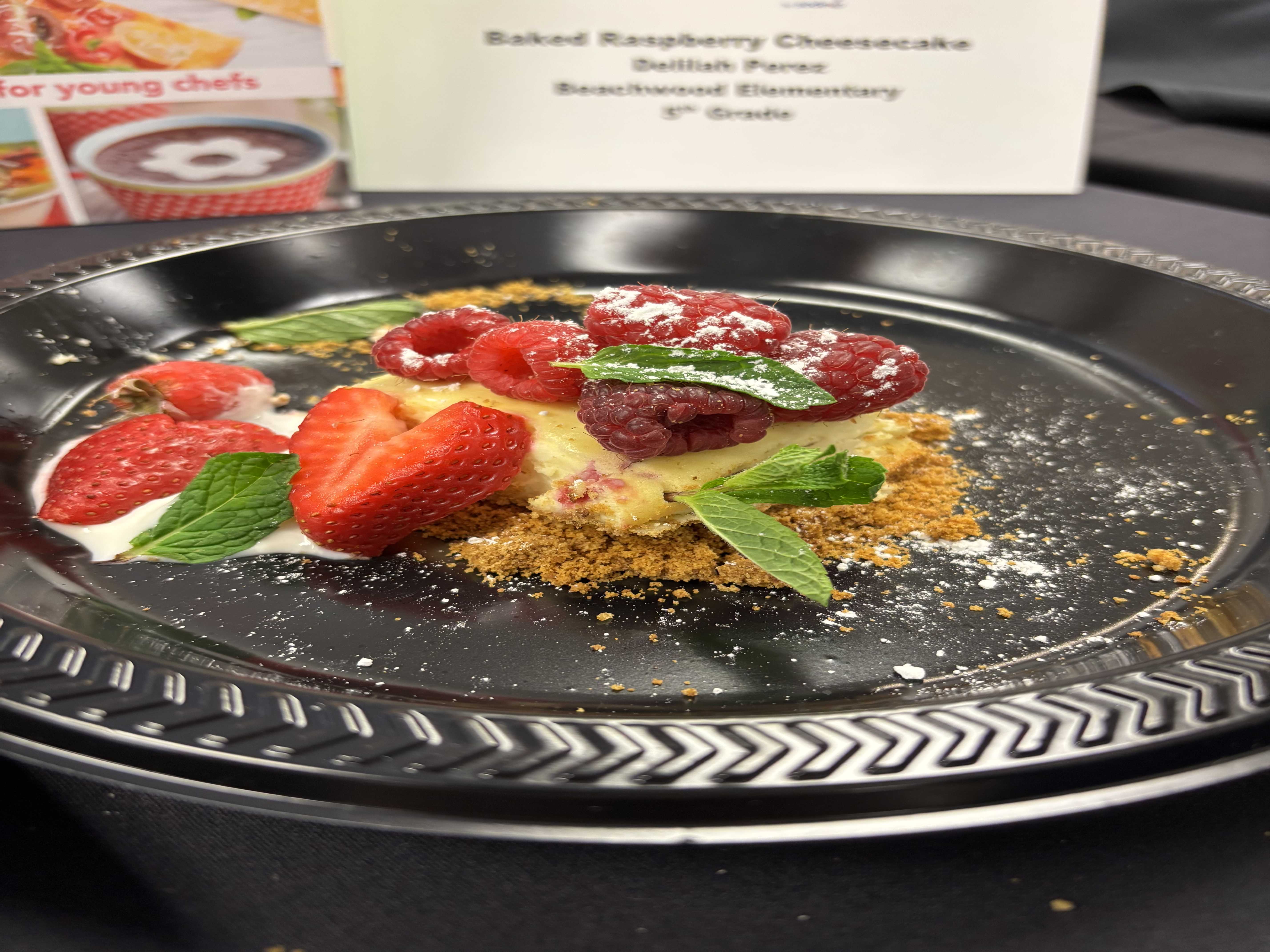 An image of a white cheesecake dessert with strawberries, raspberries, mint leaves and a crumbly tan food on top. The food is on a black plastic plate that is on top of a black table.